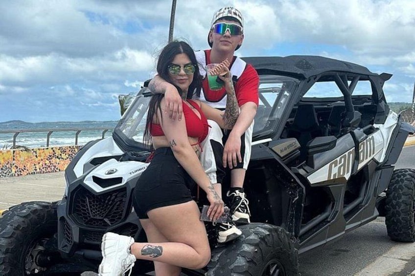 Two people sitting on a black off-road vehicle with the word 'Can-am' on the side, parked on a paved area with a cloudy sky in the background. 3