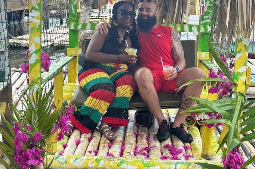 Two people sitting on a bench with colorful striped fabric, surrounded by greenery and flowers. 3