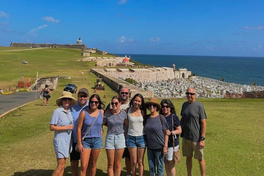 A group of people standing on a grassy area with a historic fort and ocean in the background. 2