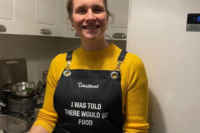 Una persona indossa un maglione giallo e un grembiule nero con scritte, in piedi in una cucina con pensili e un fornello. 4