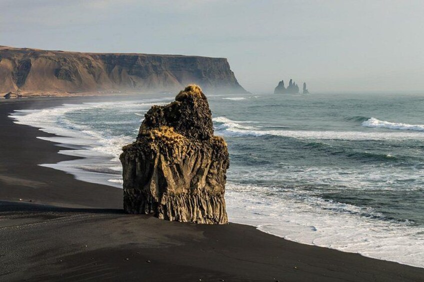 A rocky formation on a beach with waves crashing nearby and a cliff in the background. 3