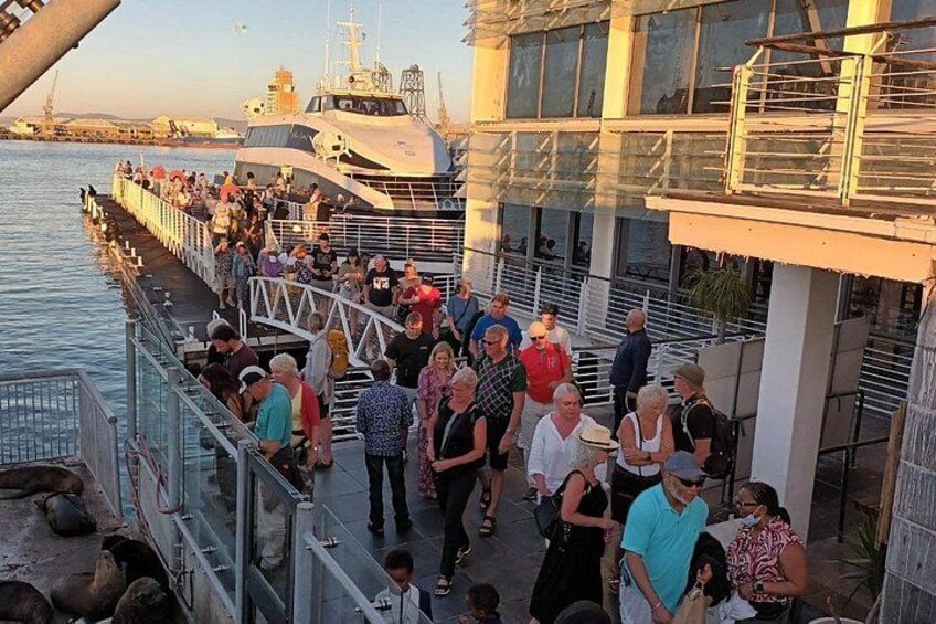 Ein belebter Steg mit Menschen, die darauf laufen und stehen, ein großes Schiff im Hintergrund und ein Gebäude mit Glasfassade. 4