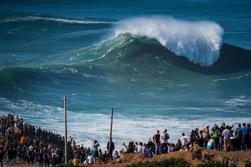 A large wave in the ocean with a crowd of people watching from the shore. 1