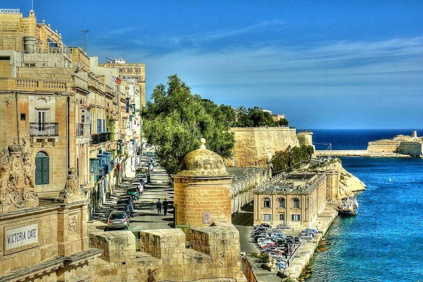 A coastal cityscape with historic buildings, a waterfront promenade, and a clear blue sea. 3