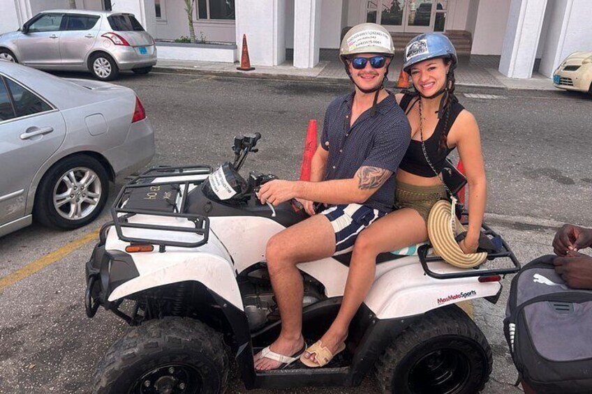 A couple on a white ATV with a helmet and a bag, parked in a lot with buildings and cars in the background. 2
