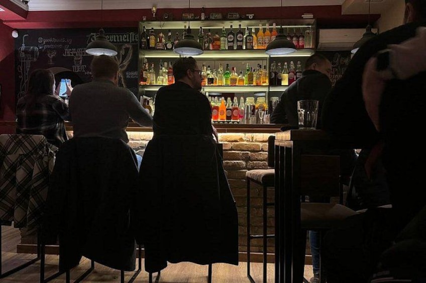 A bar with people standing at the counter, a bartender, and shelves stocked with bottles. 3