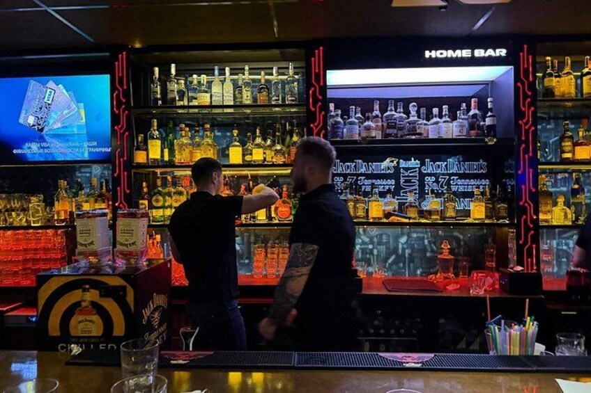 A bar with shelves of liquor bottles, a television screen, and two people standing behind the bar. 4