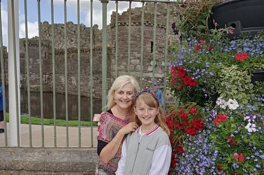 Twee personen staan voor een stenen muur met een metalen hekwerk, en een bloemenmuur aan de rechterkant. 1