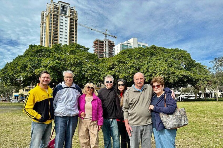 Un groupe de personnes se tient dans un parc, avec un grand arbre et des bâtiments en arrière-plan. 2