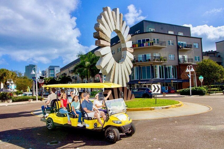 Un carrito de golf amarillo con pasajeros, una gran escultura circular y edificios modernos al fondo. 5