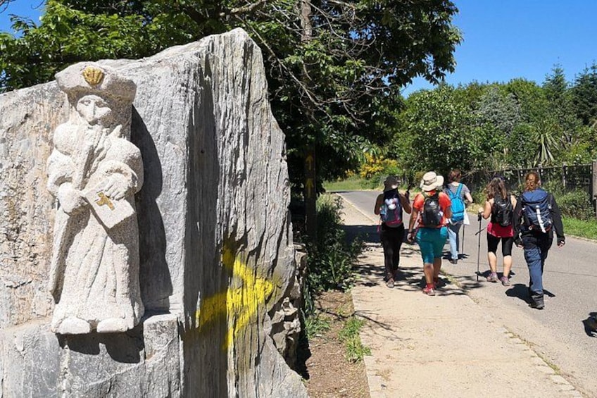 Eine Steinstatue einer Figur mit einem Kreuz, eine Gruppe Wanderer auf einem Pfad und eine Straße mit Bäumen im Hintergrund. 3