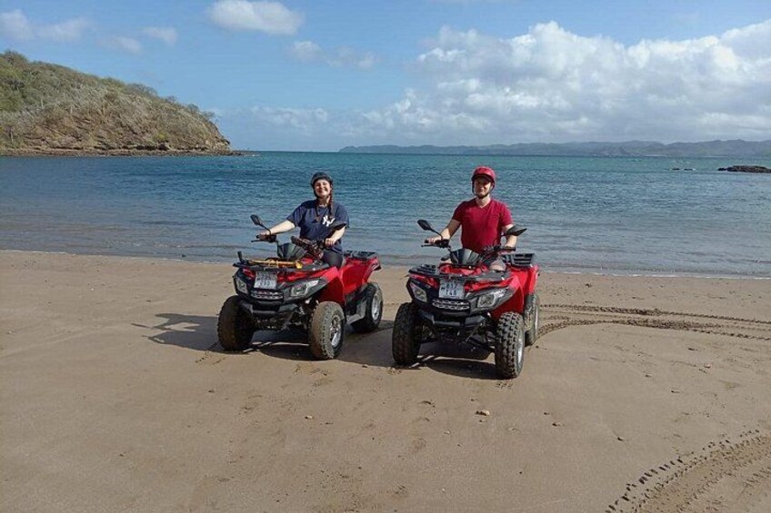 Dos personas en cuatrimotos rojas sobre una playa arenosa, con un cuerpo de agua y montañas distantes al fondo. 1