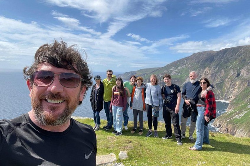 A group of people standing on a grassy hill with a mountainous landscape and the ocean in the background. 1