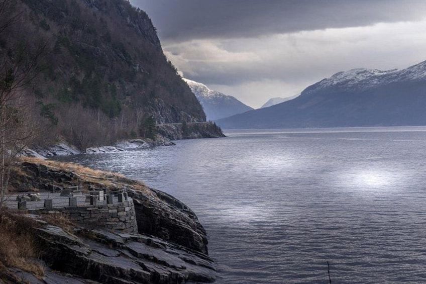 A coastal landscape with a rocky shoreline, a stone structure, and snow-capped mountains in the background. 4
