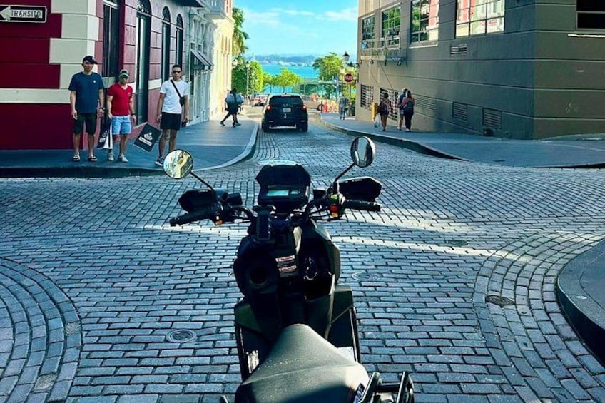 En kullerstensgata med en parkerad motorcykel, byggnader och fotgängare. 3