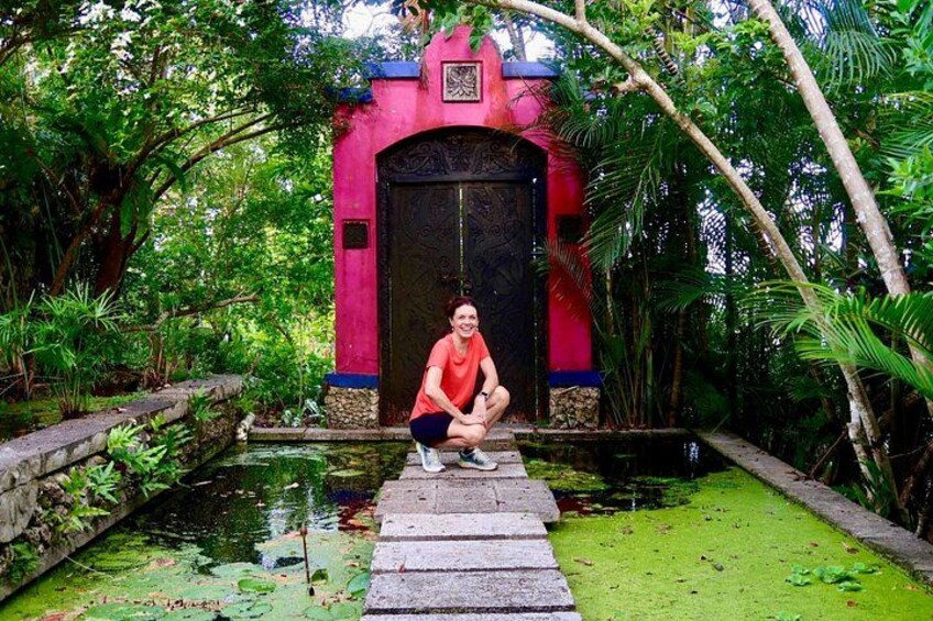 Eine Person kauert vor einem leuchtend pinkfarbenen Tor mit einer schwarzen Tür, umgeben von üppigem Grün und einem kleinen Teich. 2