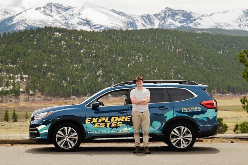 A person standing next to a blue SUV with 'EXPLORE ESTES' written on it, parked on a road with a mountainous landscape in the background. 2