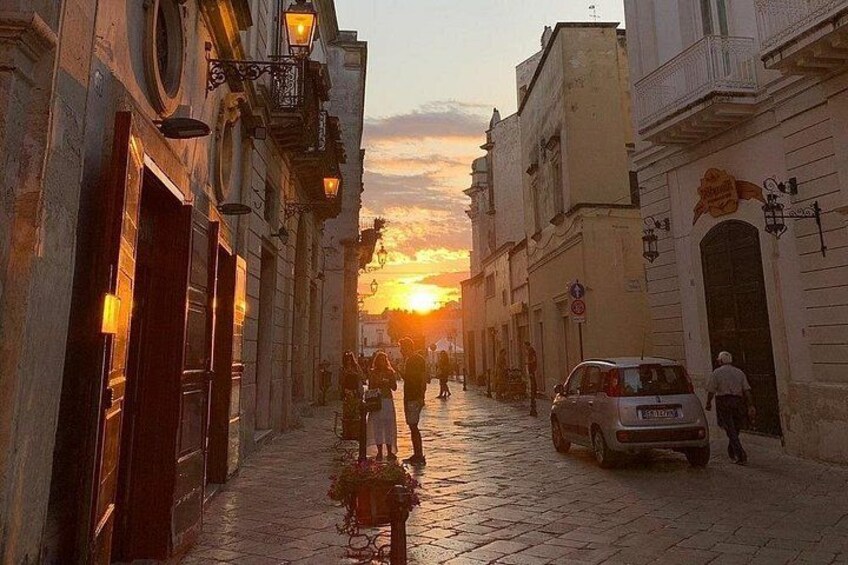 A cobblestone street at sunset with buildings on either side, a parked car, and a person walking. 2