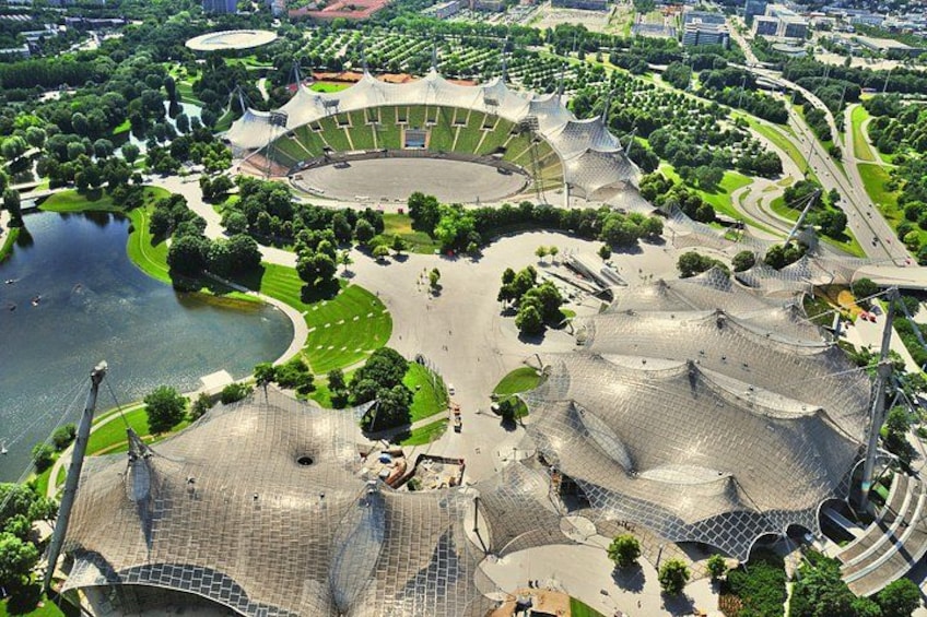 An aerial view of a modern architectural complex with a central stadium, surrounding green spaces, and a body of water. 5