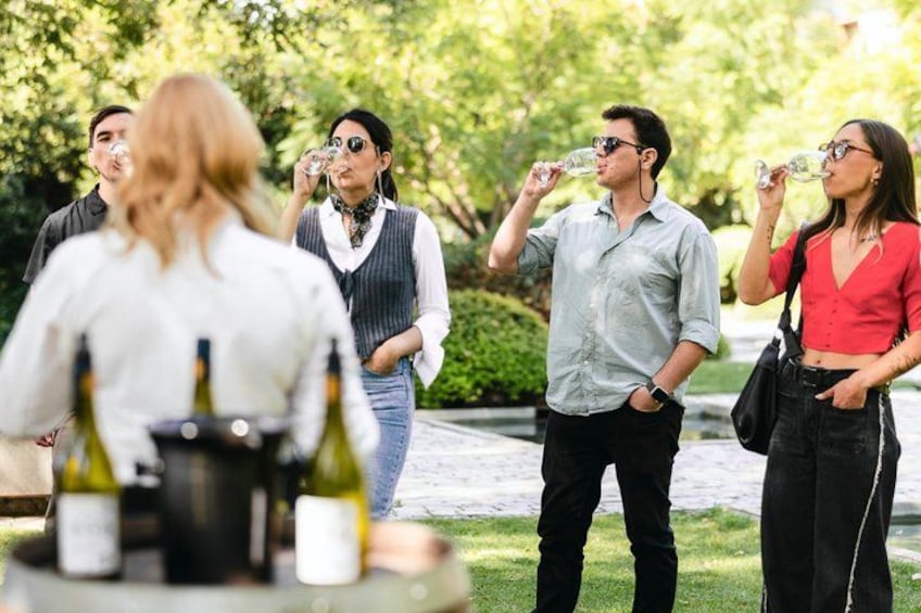 A group of people outdoors, some holding wine glasses, with bottles on a table in the foreground. 4