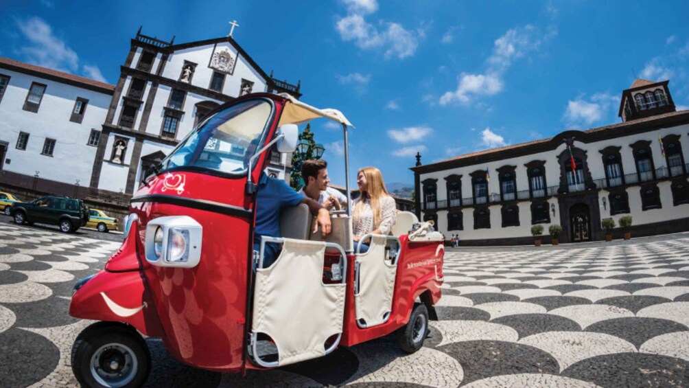 A red and white tuk-tuk with passengers, parked on a patterned pavement with historic buildings in the background. 5