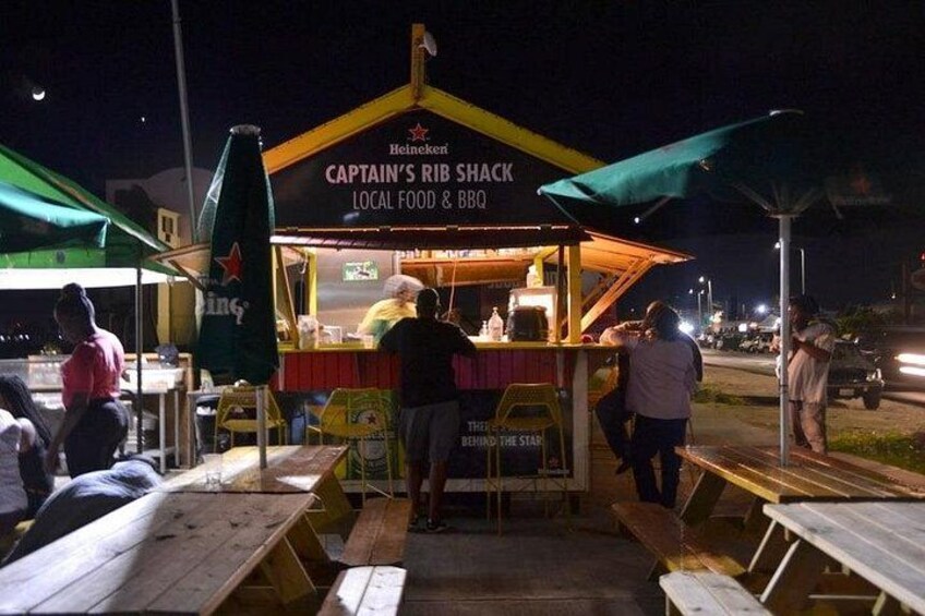 A food stand named 'Captain's Rib Shack' with a sign indicating it serves local food and BBQ. There are tables and chairs around the stand, and people are gathered around. 5