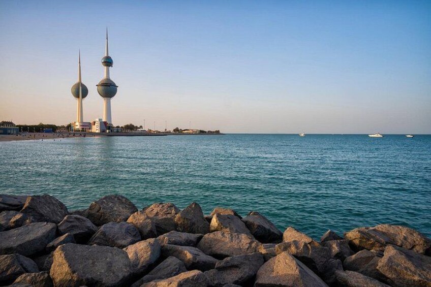A coastal scene with large rocks in the foreground, a calm sea, and two prominent towers in the background. 2