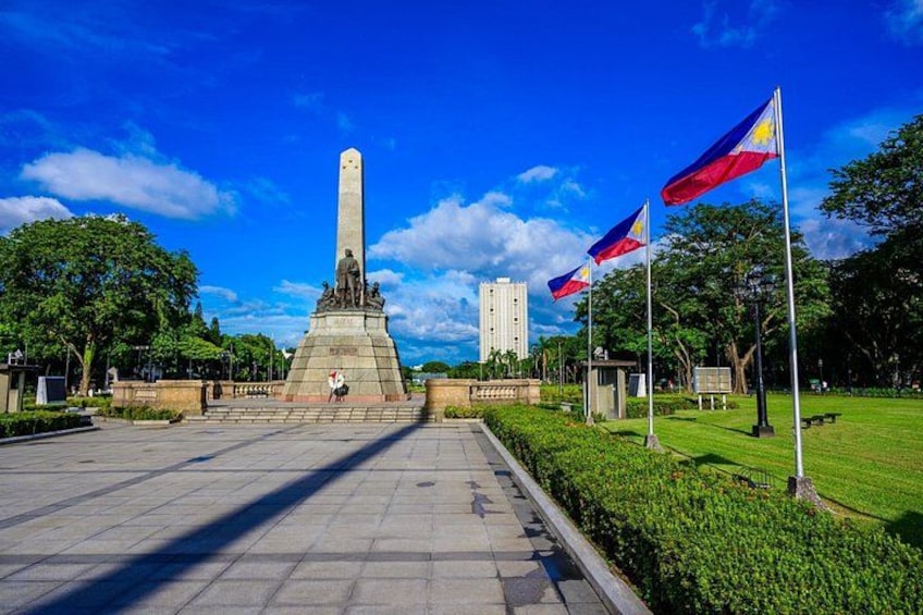 A monument with a tall obelisk, surrounded by a park with grass, trees, and a clear blue sky. 2