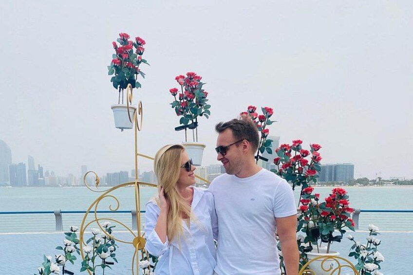A couple standing by a decorative metal structure with flowers, overlooking a body of water with a city skyline in the background. 3