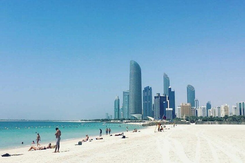 A beach with people sunbathing and swimming, a clear sky, and a city skyline with tall buildings in the background. 2