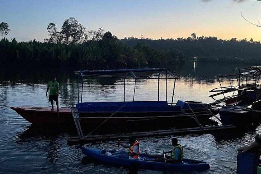 A calm river with boats docked, a person standing on a boat, and another person in a kayak. 4