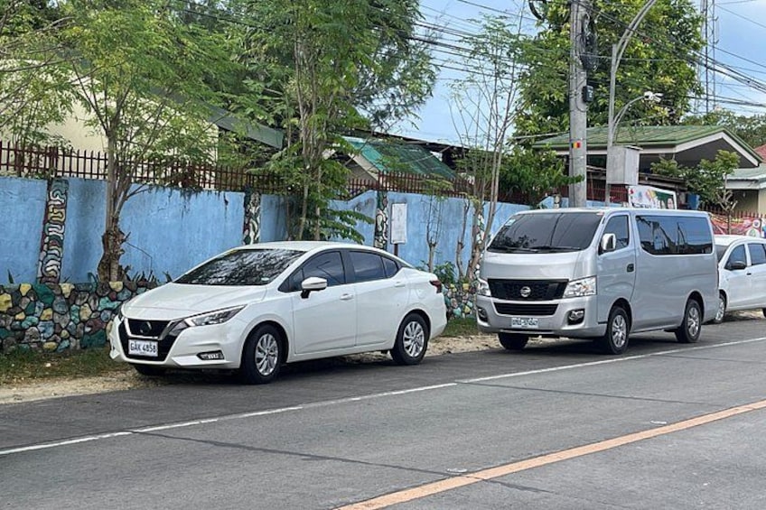 A white car parked on the side of a road, a silver van, and a blue wall with a stone pattern. 2