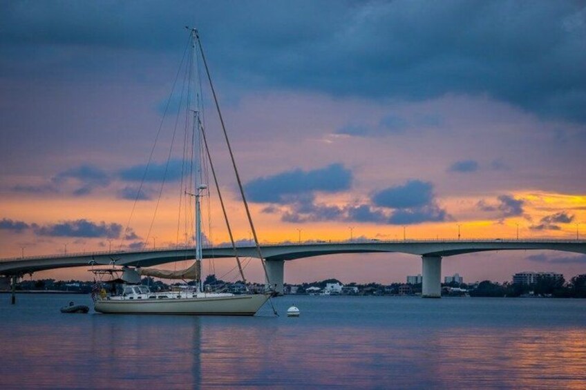Un velero sobre aguas tranquilas, un puente al fondo y un cielo al atardecer. 1