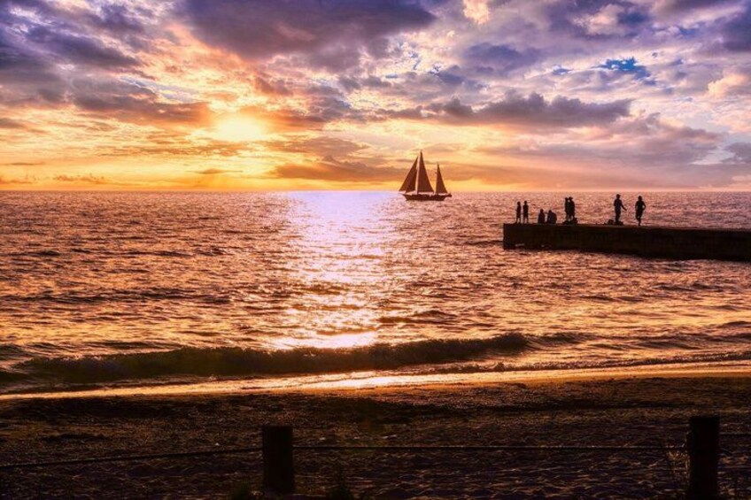 Atardecer sobre el océano con un velero y gente en un muelle. 4