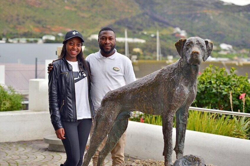Ein Paar steht neben einer bronzenen Hundstatue in einem Park mit See und Bergen im Hintergrund. 2