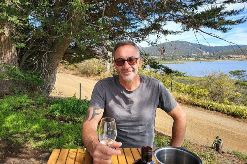 A man sitting at a wooden table outdoors with a glass of wine, a metal bowl, and a bottle. 2