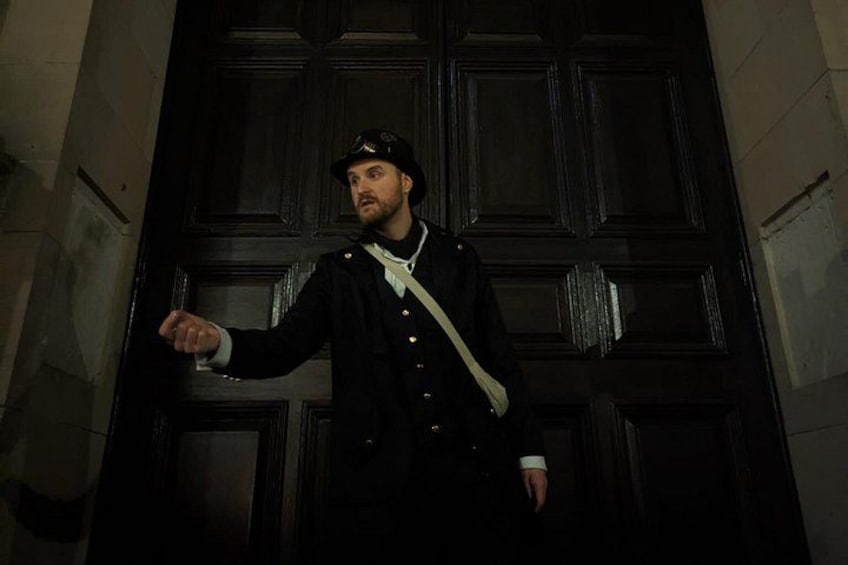 A person in a dark coat and hat standing in front of a dark wooden door. 2