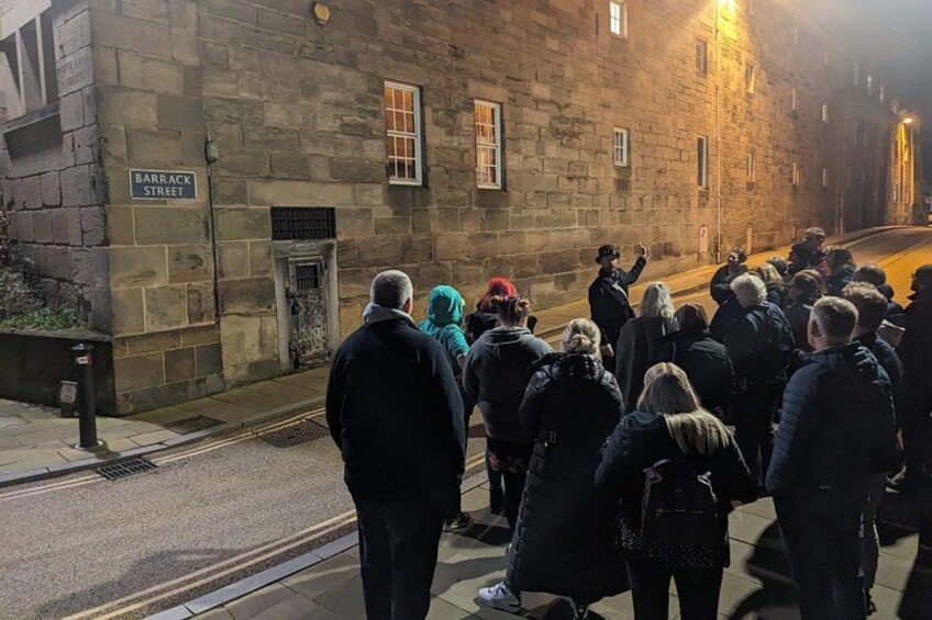 A group of people standing on a street in front of a stone building with a sign that reads 'Barrack Street.' 1
