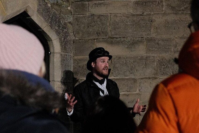 A person in a black hat and coat gesturing in front of a stone wall. 4