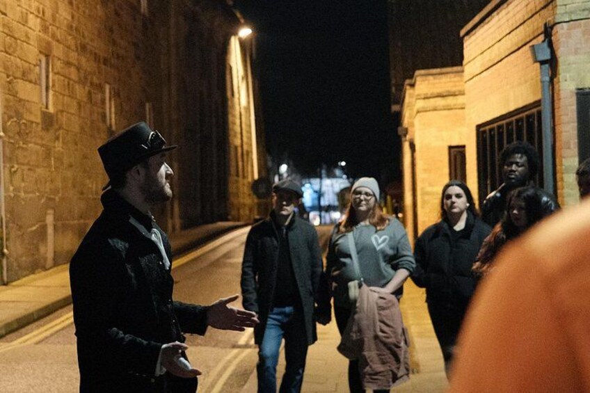 A nighttime street scene with a person in a top hat and coat gesturing to a group of people. 3