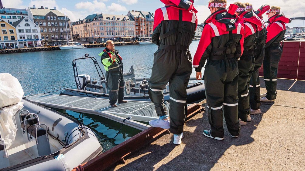 Un groupe de personnes vêtues d’uniformes rouges et noirs se tient sur un quai, au bord d’une étendue d’eau, avec des bâtiments en arrière-plan. 2