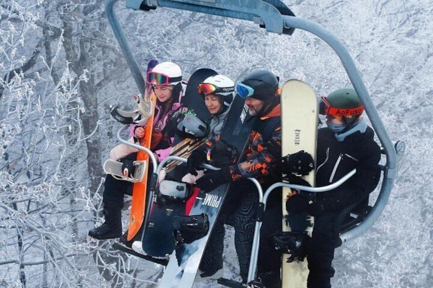 A group of people on a ski lift, holding snowboards, with snow-covered trees in the background. 4