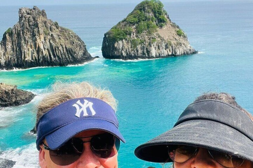 A person wearing a baseball cap and another person with a hat, overlooking a coastal landscape with rocky formations and turquoise waters. 4