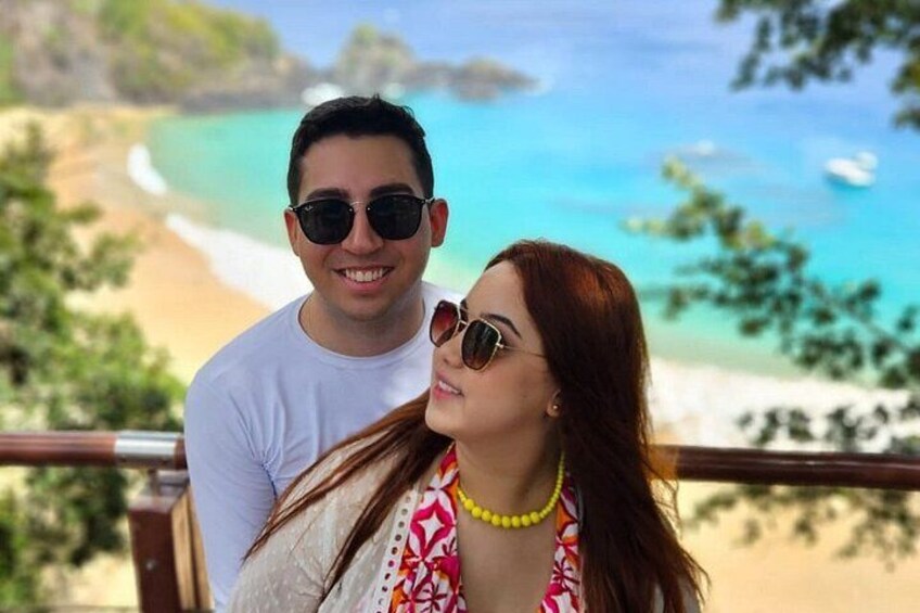 Un couple se tient sur un balcon donnant sur une plage, avec une eau turquoise cristalline et une rive sablonneuse. 2