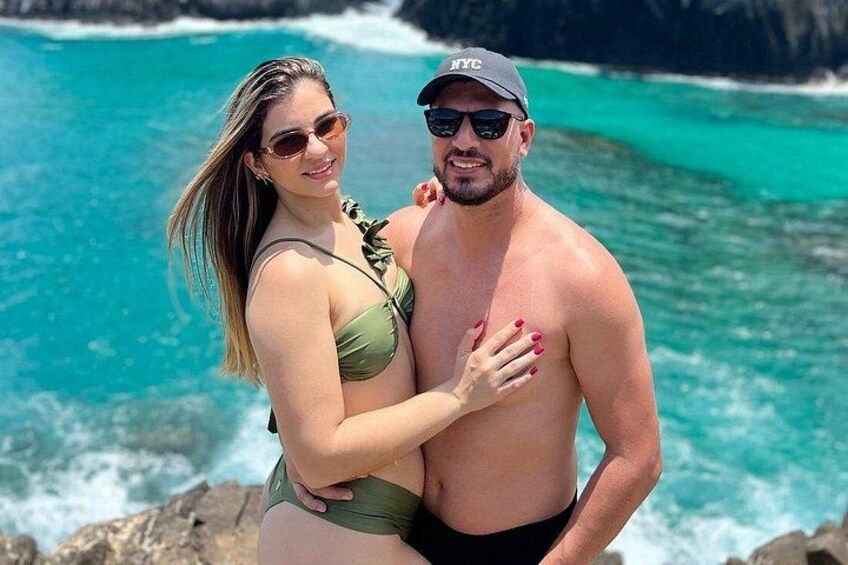 A couple standing by a turquoise ocean with rocky cliffs in the background. 4