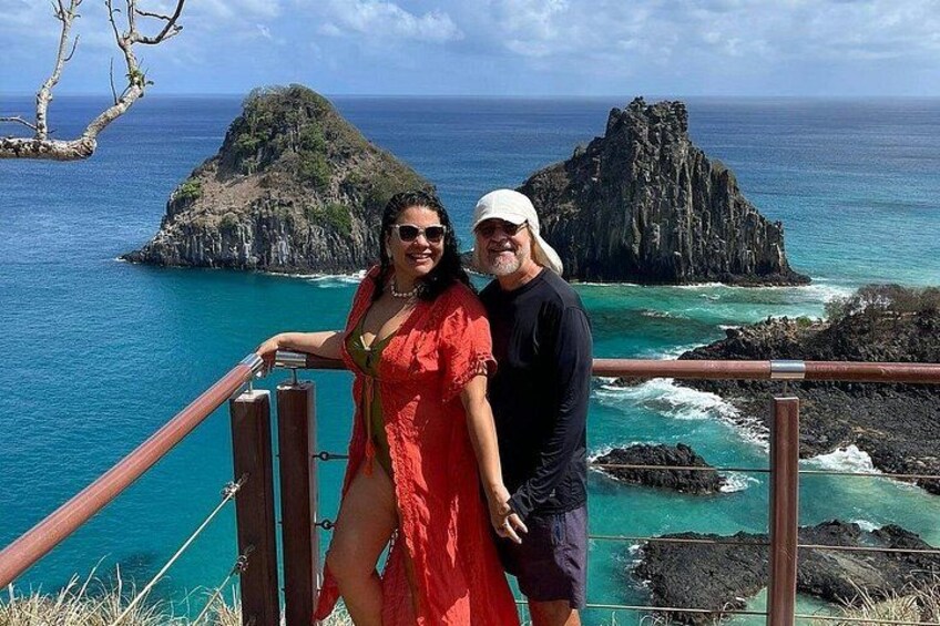 A couple standing on a wooden railing overlooking a coastal landscape with rocky outcrops and turquoise waters. 3