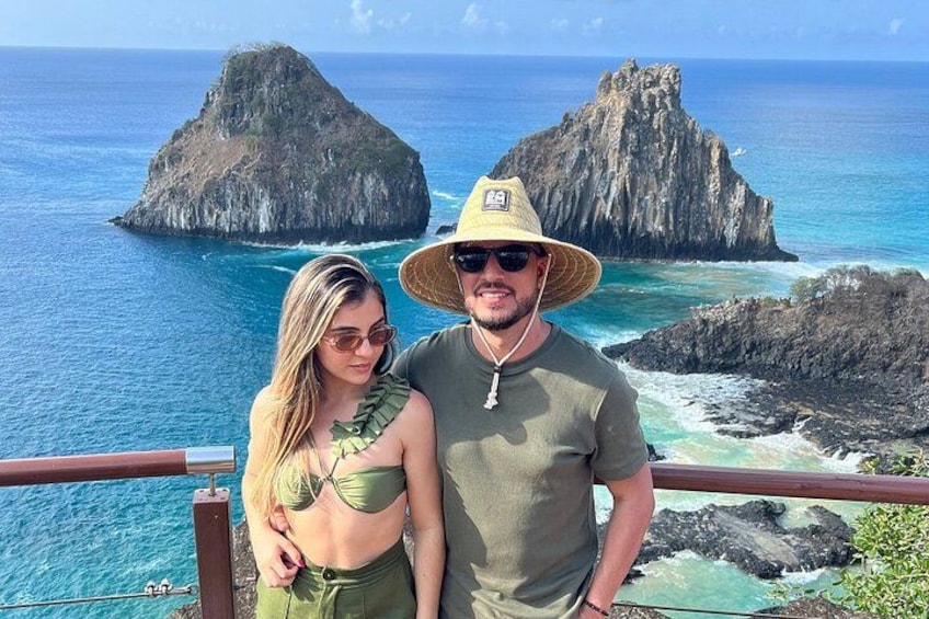 Un couple se tient près d’une rambarde, d’où l’on peut admirer un littoral rocheux et des eaux d’un bleu limpide. 1