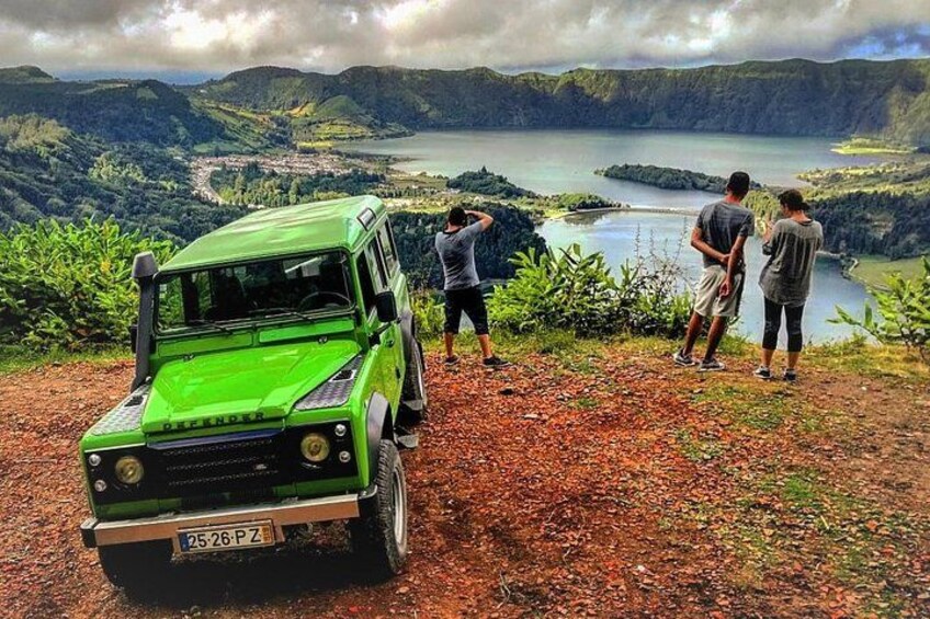 Un fuoristrada verde parcheggiato su un sentiero sterrato con vista su un lago, vicino al quale si trovano persone. 1