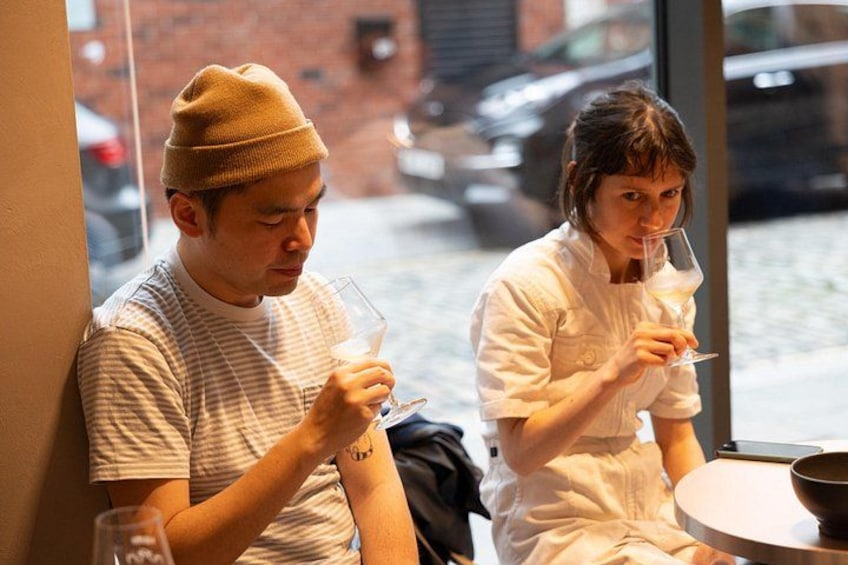 Two people sitting at a table with a glass of wine, a bowl, and a window with a view of the street outside. 3
