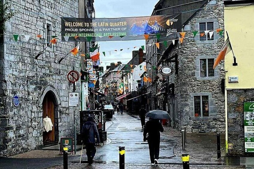 A wet street with stone buildings, a sign welcoming to the Latin Quarter, and flags. 3